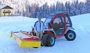 Ein Mann steht neben einem Reform Metrac G5 X Fahrzeug im Schnee vor einem gelben Haus und schneebedeckten Bäumen.