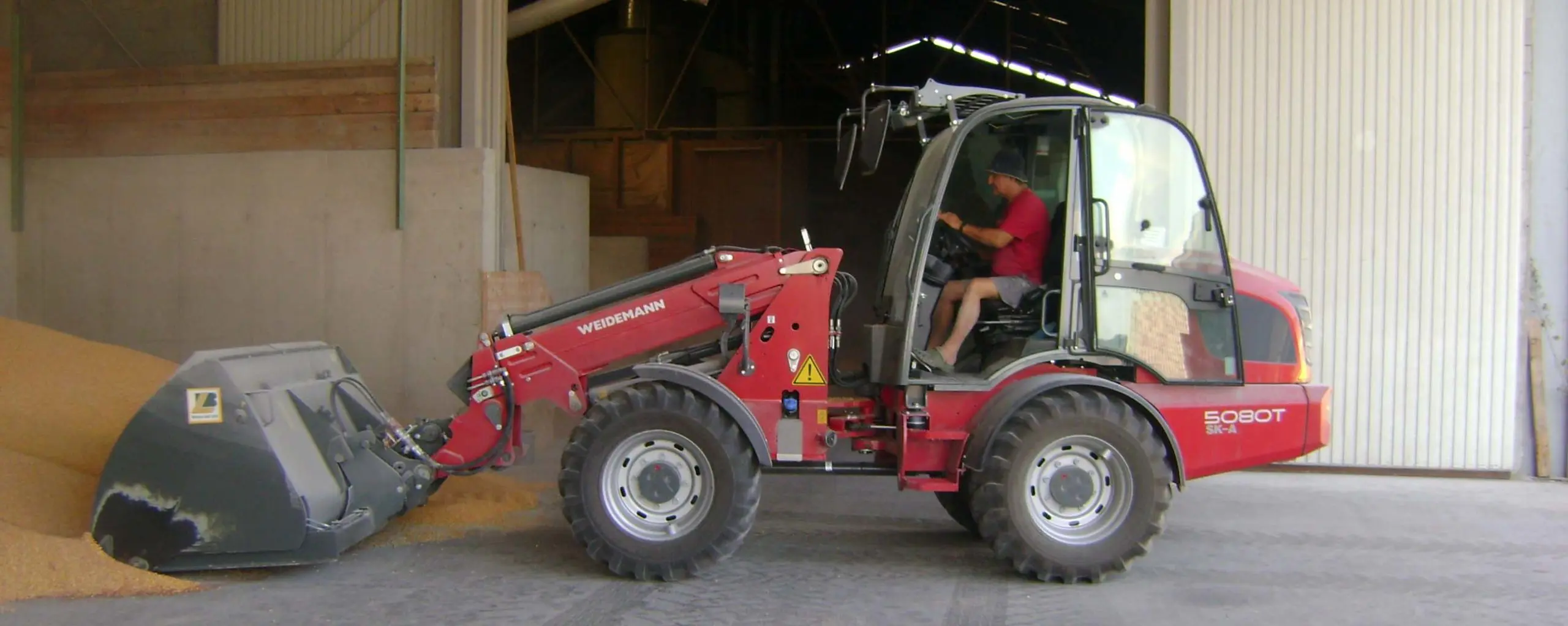 Ein Mann bedient einen roten Weidemann Radlader 5080T SK-A, um Getreide in einer Lagerhalle zu bewegen.