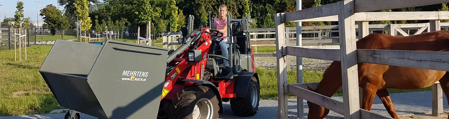 Person fährt auf einem roten Mehrtens-Mauch-Traktor vor einem Pferd in einem eingezäunten Bereich.