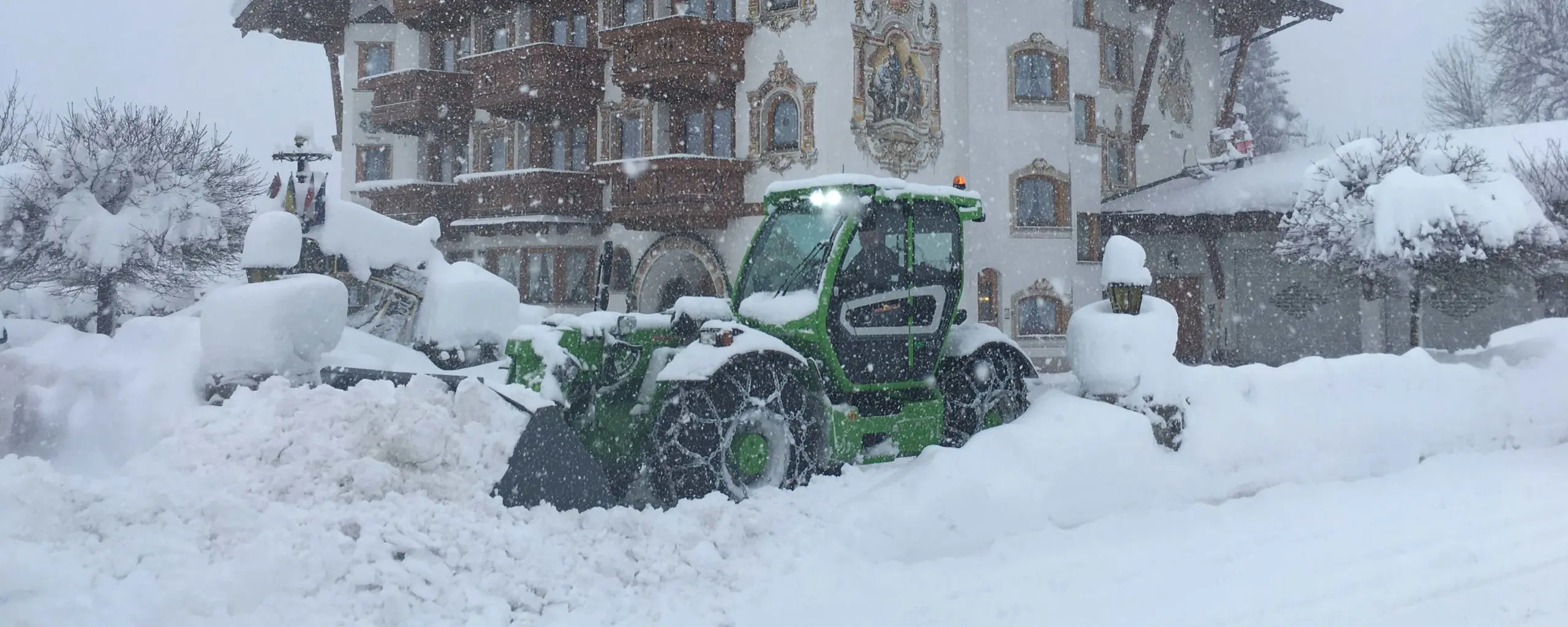 Ein grüner Traktor schaufelt Schnee vor einem verschneiten Gebäude.