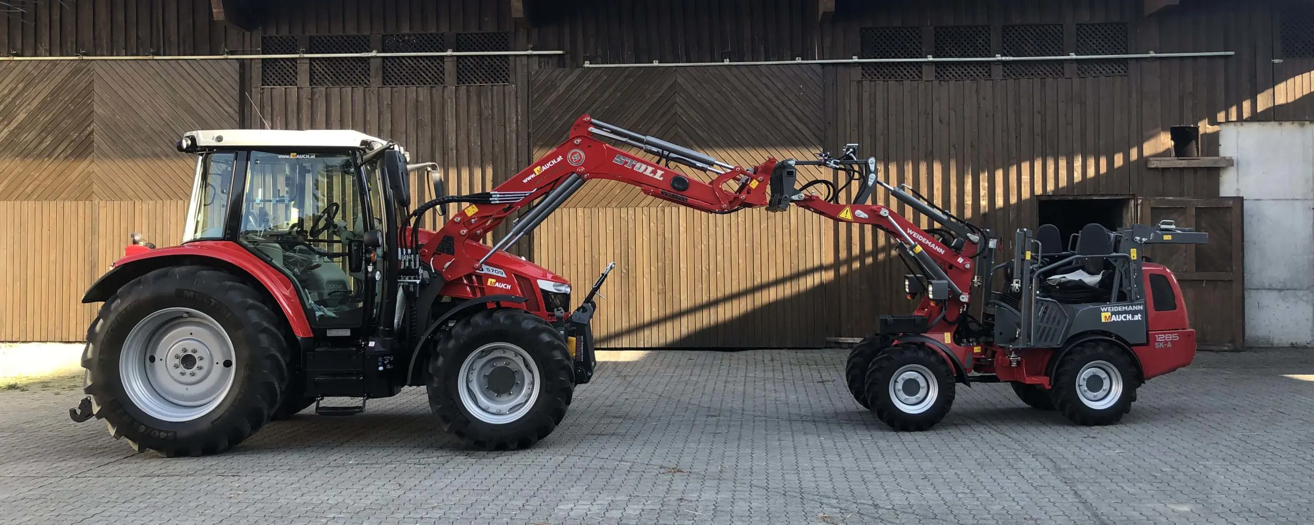 Ein roter Traktor und ein roter Weidemann-Lader stehen nebeneinander vor einer Holzwand.