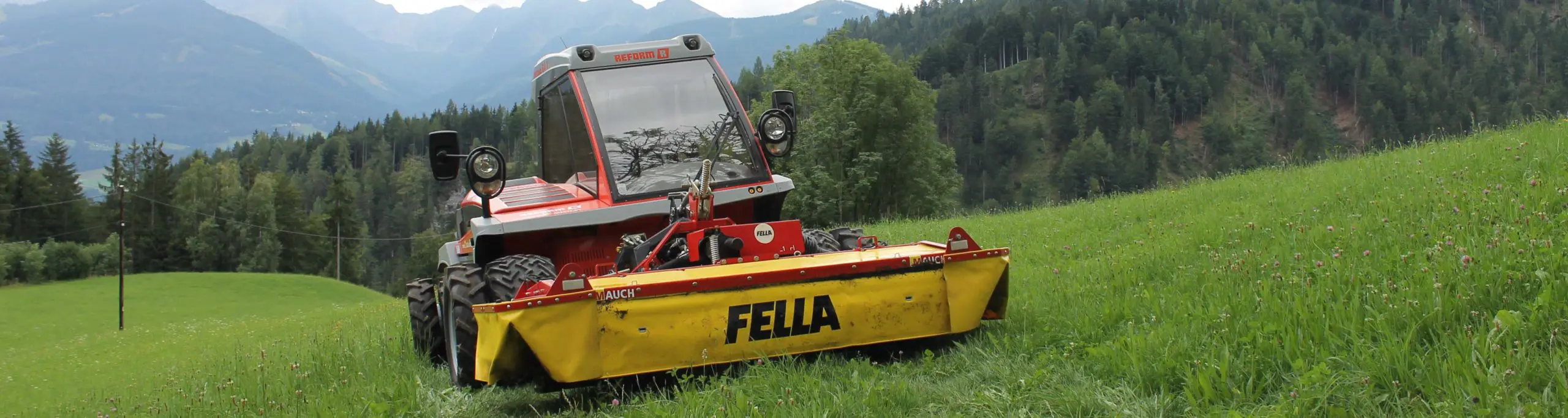 Ein roter Reform R Traktor mit Fella-Mähwerk auf einer grünen Wiese vor Berglandschaft.