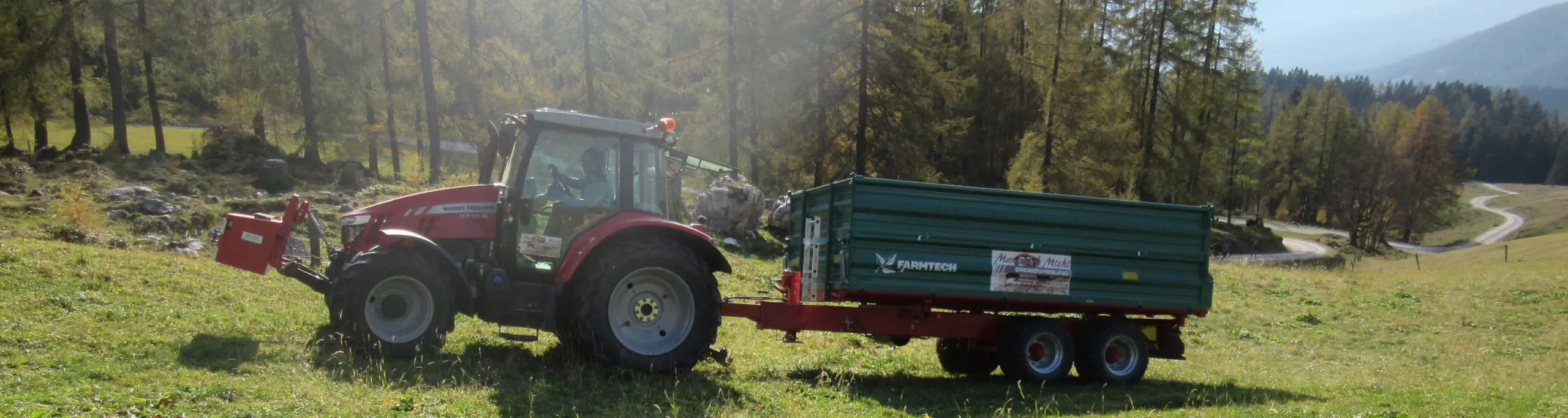 Ein roter Massey Ferguson Traktor zieht einen grünen Farmtech Anhänger auf einer Wiese.