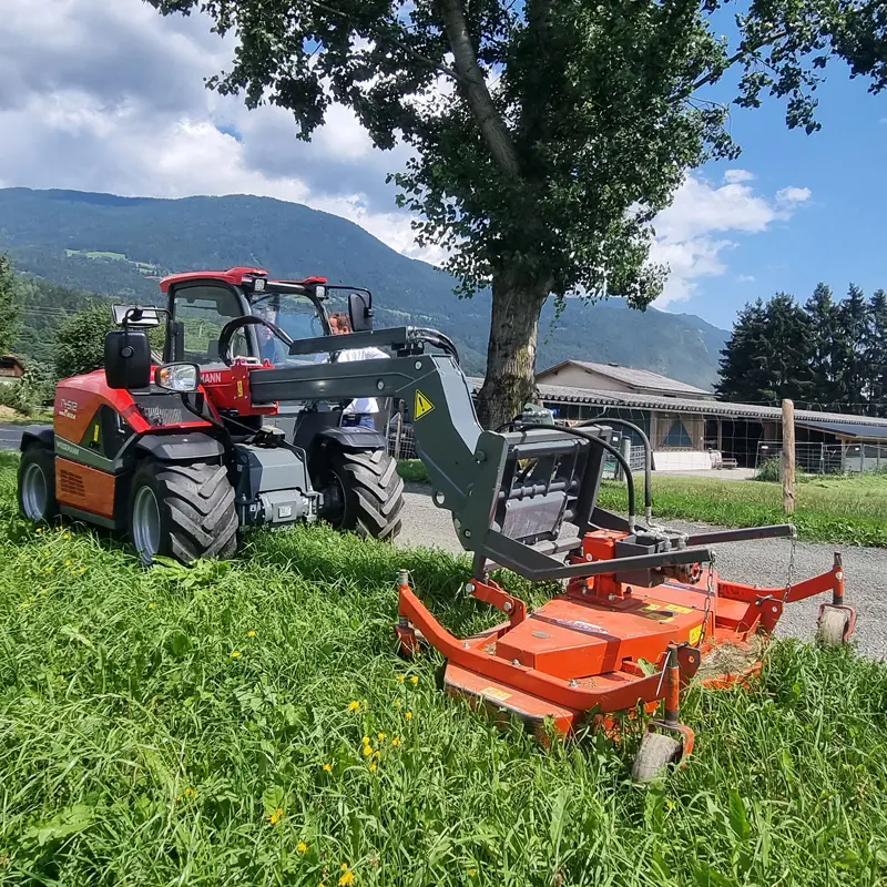 Ein Weidemann T4512 steht auf einer Wiese und hebt eine orange landwirtschaftliche Maschine an.