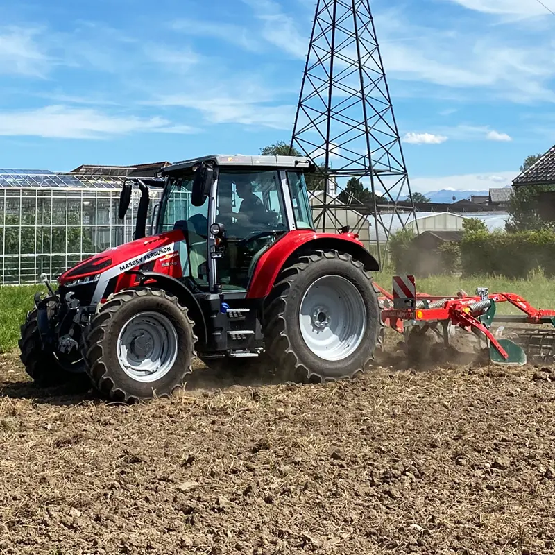 Massey Ferguson Traktor pflügt ein Feld vor einem Gewächshaus und einem Strommast.