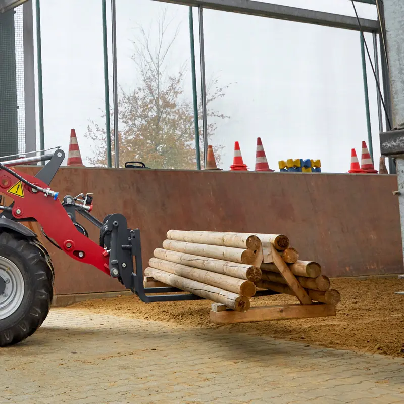 Roter Traktor mit Staplergabel hebt Holzstämme in einer Halle.