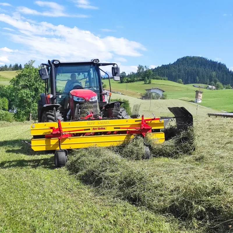 Traktor MF4708M zieht Sepp Knüsel Heuwender auf einem Feld mit grüner Landschaft im Hintergrund.