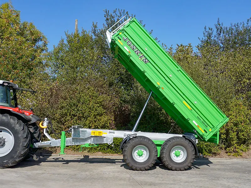 Remorcă basculantă Joskin Delta-CAP verde cu două axe, cuplată în lateralul tractorului, cu benă basculantă ridicată hidraulic.