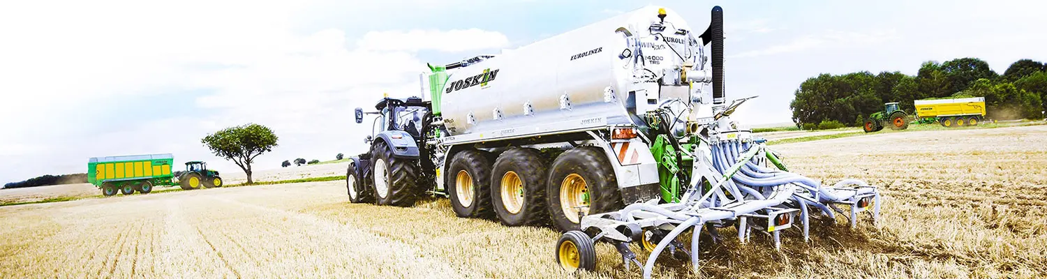Cisternă pentru nămol Joskin Euroliner în spatele unui tractor care împrăștie nămol pe un câmp de miriște, cu alte utilaje agricole în fundal