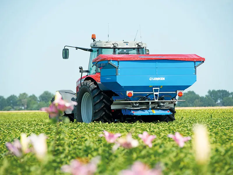 Un distribuitor de îngrășăminte Lemken Tauri atașat la un tractor traversează un câmp verde cu flori în prim-plan.
