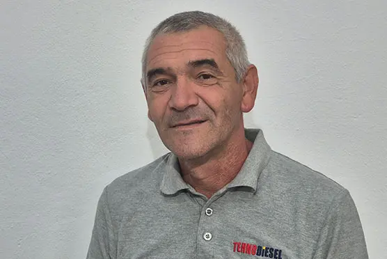 Un bărbat cu părul gri, purtând un tricou polo gri cu sigla TechnoDiesel, stă în fața unui perete alb.