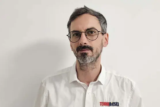 Bărbat cu ochelari și barbă, într-o cămașă albă cu sigla TEKNDIESEL, în fața unui perete alb