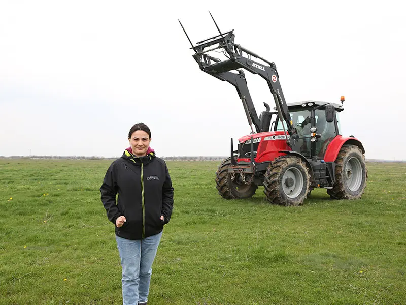 Femeie îmbrăcată în jachetă CODREA stă într-o pajiște în fața unui tractor Mauch roșu cu încărcător frontal STEYR STOLL
