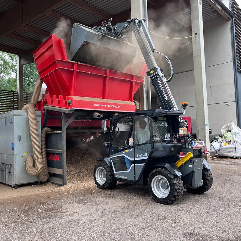 Weidemann T4512 Teleskoplader von Mauch leert Holzmaterial in große rote ZAVA‑Recyclinganlage unter Hallendach