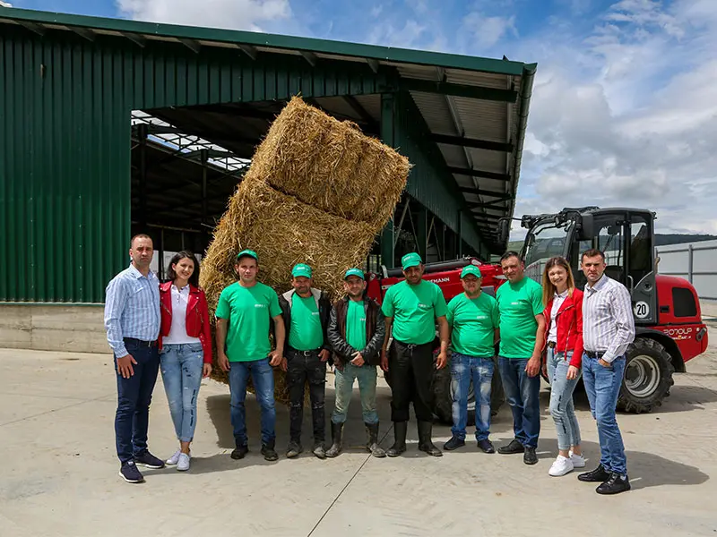 Grup de fermieri în fața sălii, lângă el un încărcător agricol roșu cu un balot de fân ridicat, echipa privind spre cameră