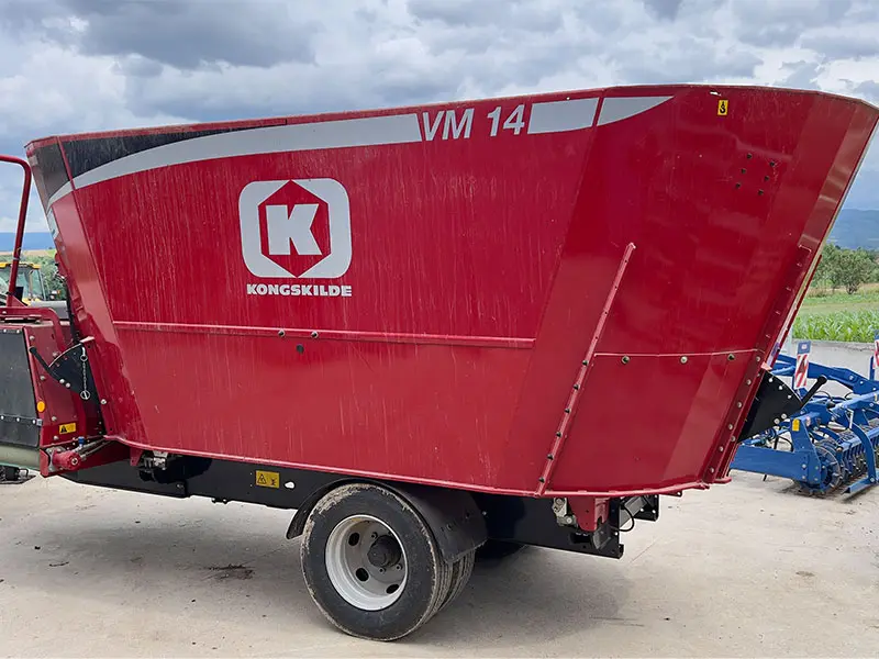 Crvena prikolica za miješanje stočne hrane Kongskilde VM 14, parkirana bočno u dvorištu, betonirana nasuprot oblačnom nebu.