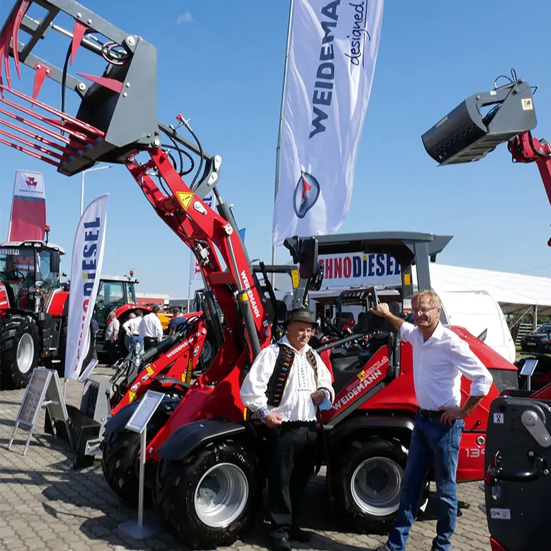 Doi bărbați stau în fața unui încărcător frontal Weidemann roșu la o expoziție de utilaje agricole, cu steaguri în fundal.