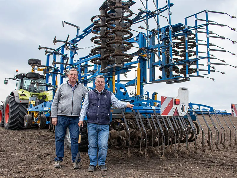 Doi bărbați stau în fața unei mașini mari de arat, albastre; în stânga se află un tractor pe un câmp.