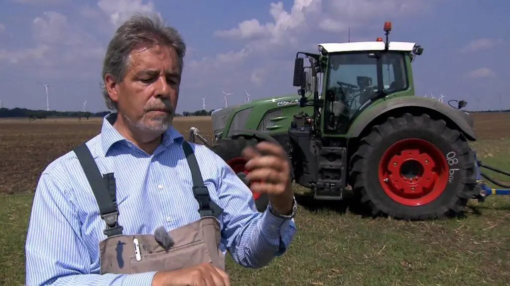 Landwirt erklärt auf Acker den Fendt-Traktor mit Variogrip-Reifen, im Hintergrund Windräder