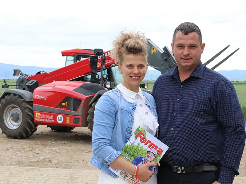 O femeie cu revista „Ferma” și un bărbat stau în fața unui încărcător telescopic Weidemann T7042 Mauch roșu pe o alee de câmp