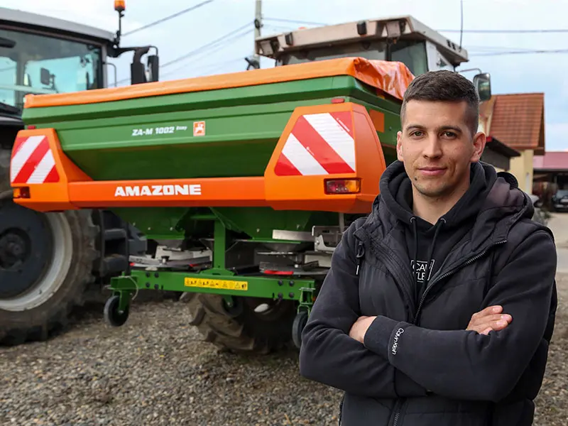 Un tânăr cu brațele încrucișate stă în fața unui distribuitor de îngrășăminte Amazone ZA-M 1002 atașat la un tractor, într-o curte.