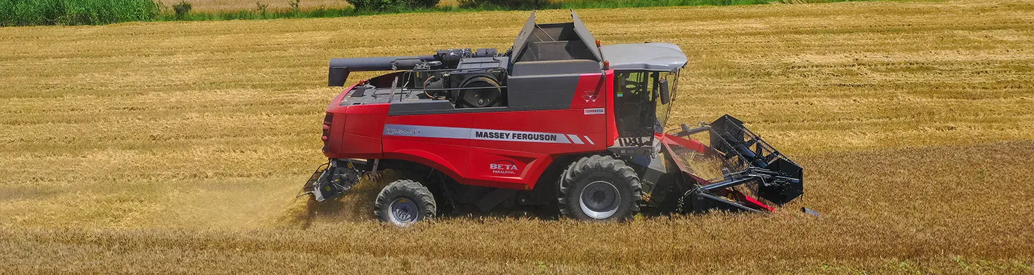 O combină de recoltat Massey Ferguson BETA roșie recoltează un câmp de cereale de la stânga la dreapta într-un peisaj deluros.