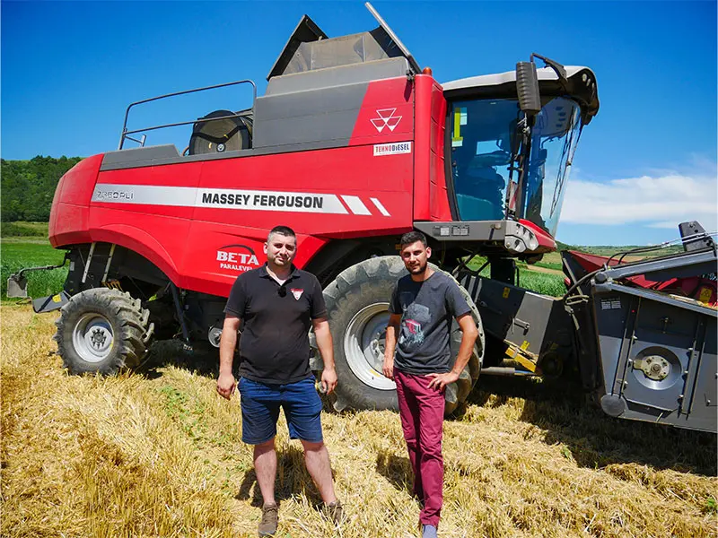 Doi bărbați stau într-un câmp de miriște în fața unei combine de recoltat Massey Ferguson roșii, cu sigla vizibilă a mărcii.