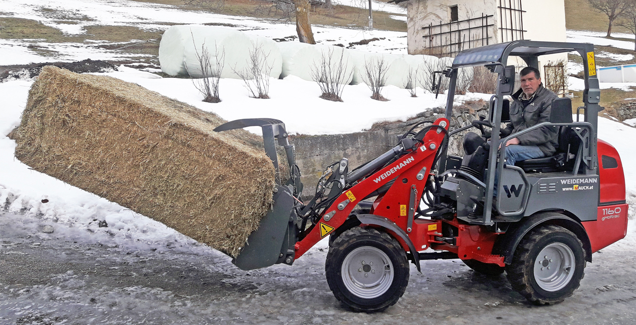 Mann fährt einen Weidemann 1160 eHoftrac Traktor mit einem Heuballen in der Schaufel im Schnee.