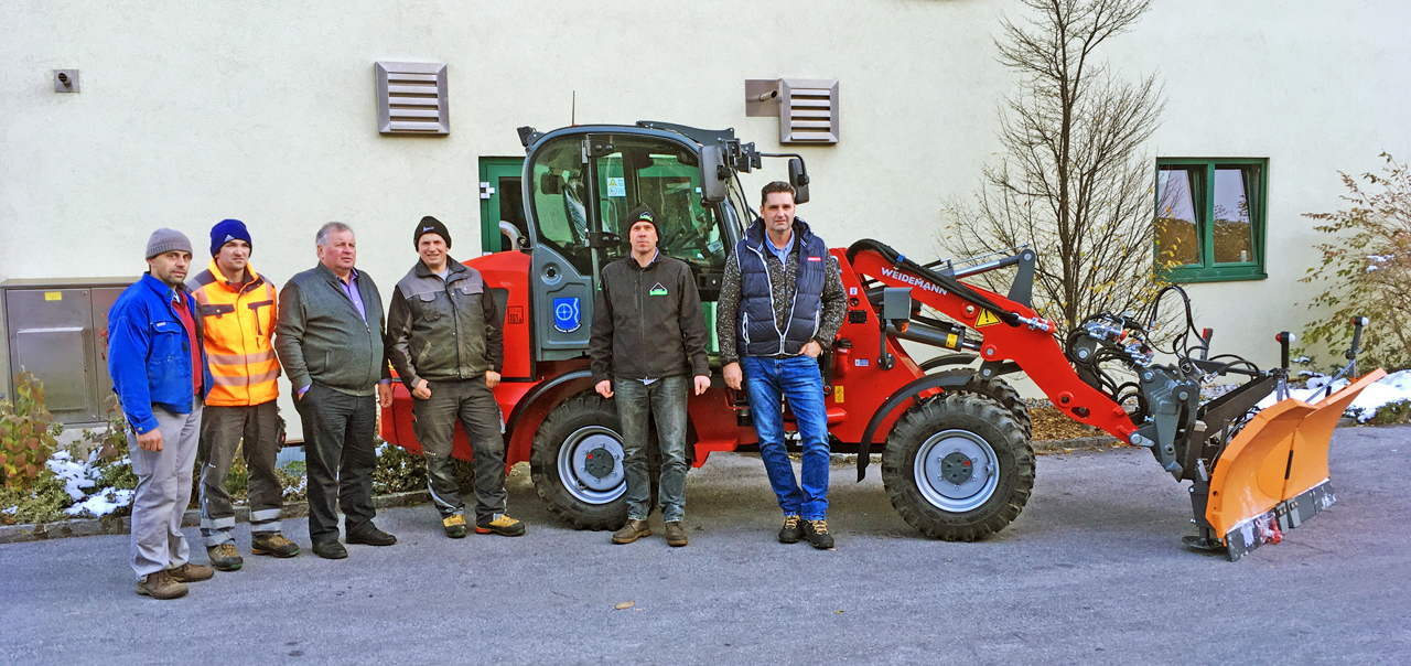 Sechs Männer stehen neben einem roten Weidemann-Traktor mit Schneepflug vor einem Gebäude.