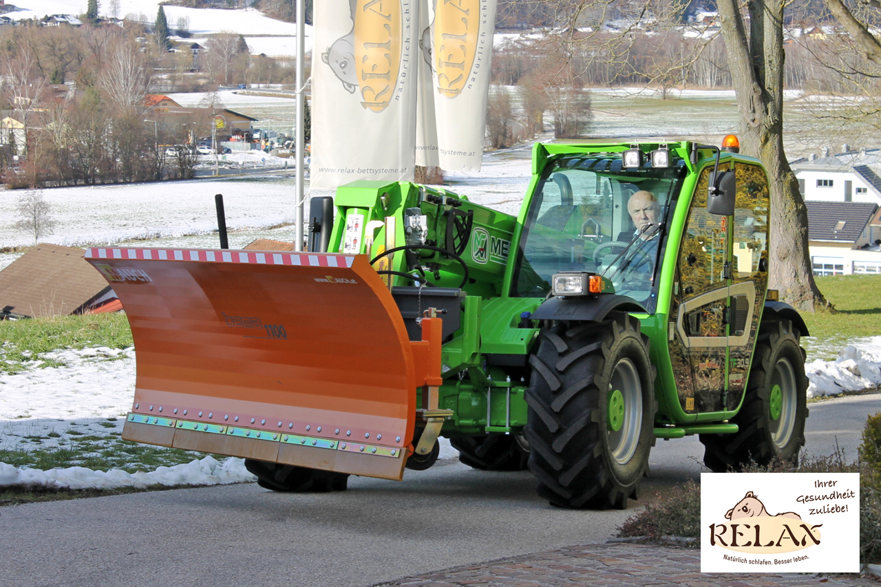 Grüner Traktor mit Schneepflug vor Relax-Flaggen auf schneebedecktem Gelände.