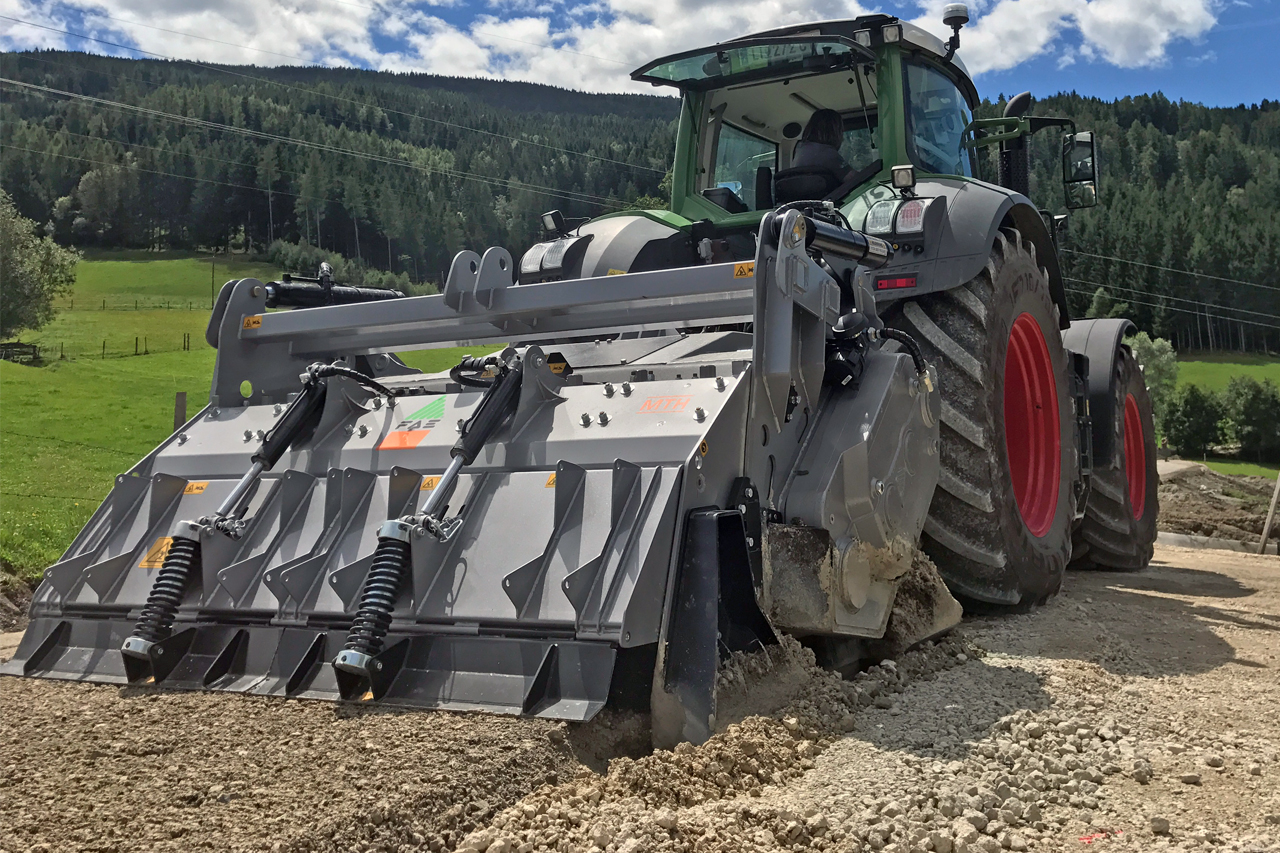 Traktor mit FAE-Gerät führt Erdarbeiten auf einem Kiesweg vor einer bewaldeten Landschaft durch.