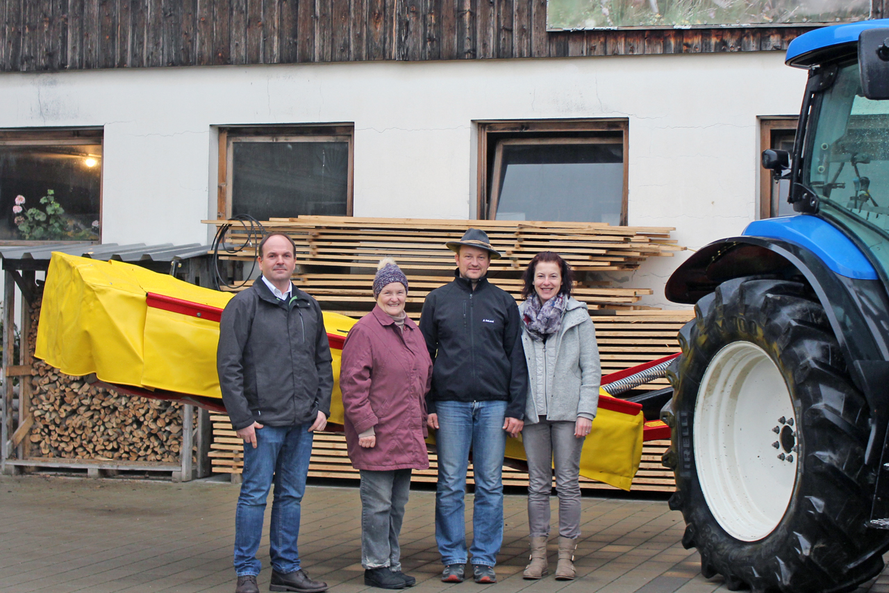 Vier Personen stehen vor einem landwirtschaftlichen Gebäude neben einem gelben Werkzeug, teilweise von einem Traktor verdeckt.