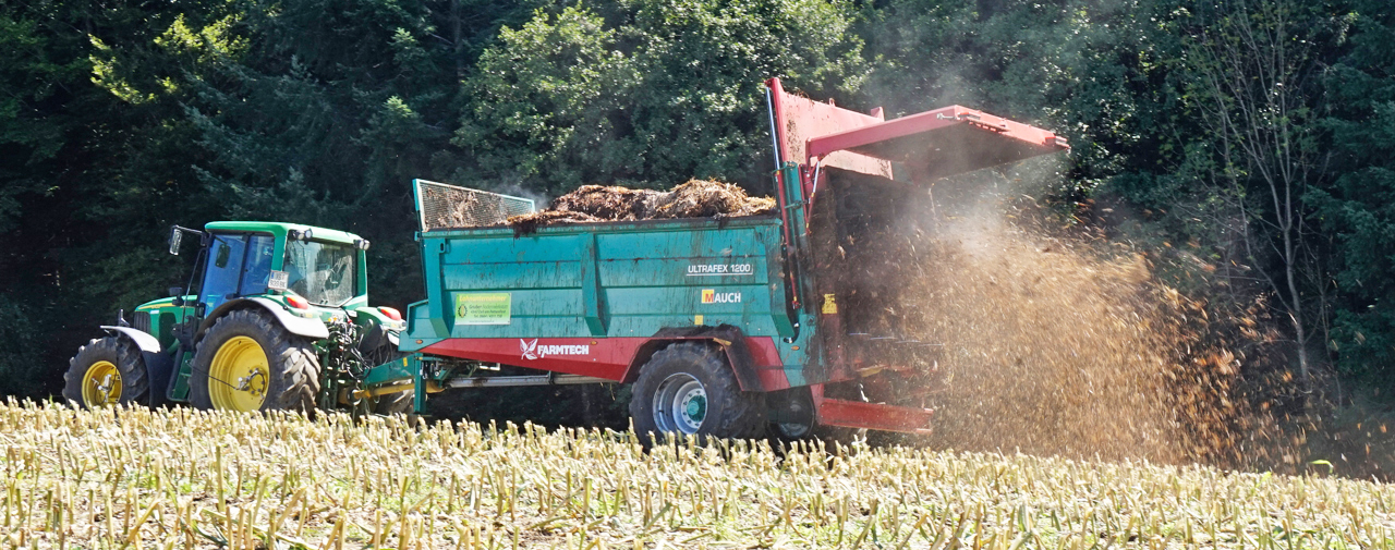 Grüner Traktor mit Farmtech Anhänger ULTRAFEX 1200 verteilt Material auf einem Feld.