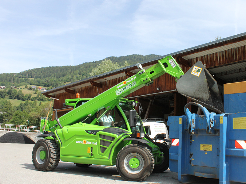 Grüner Merlo Teleskoplader P27.6 von Mauch lädt Material in einen blauen Container.