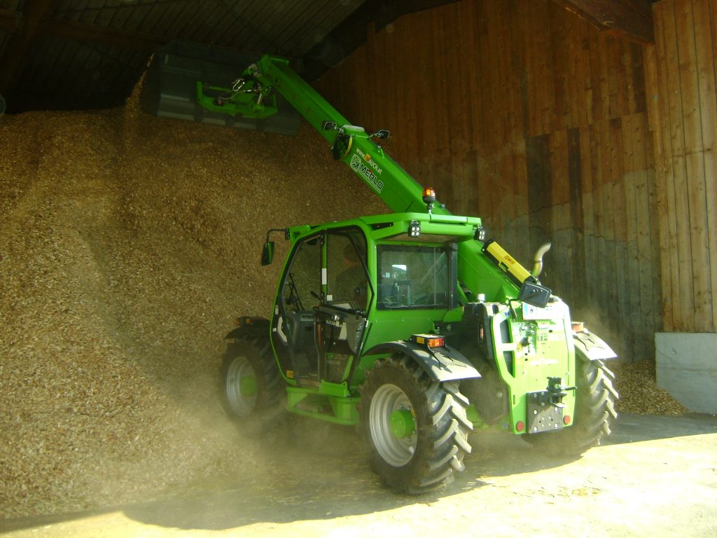 Încărcătorul telescopic Merlo verde mută așchii de lemn într-un hambar.