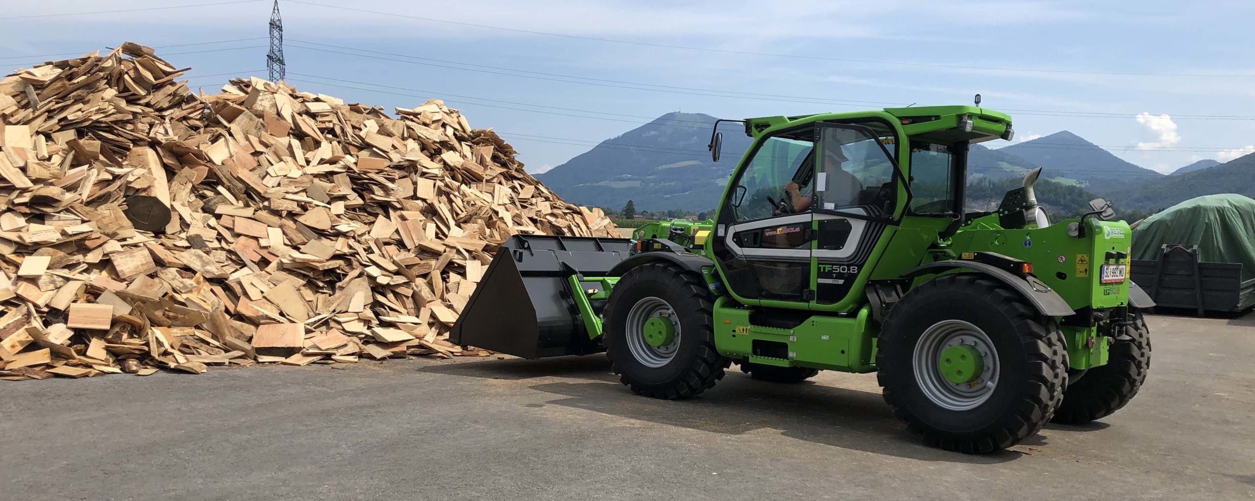 Ein grüner Merlo-Teleskoplader steht vor einem großen Holzstapel auf einem Industriegelände.