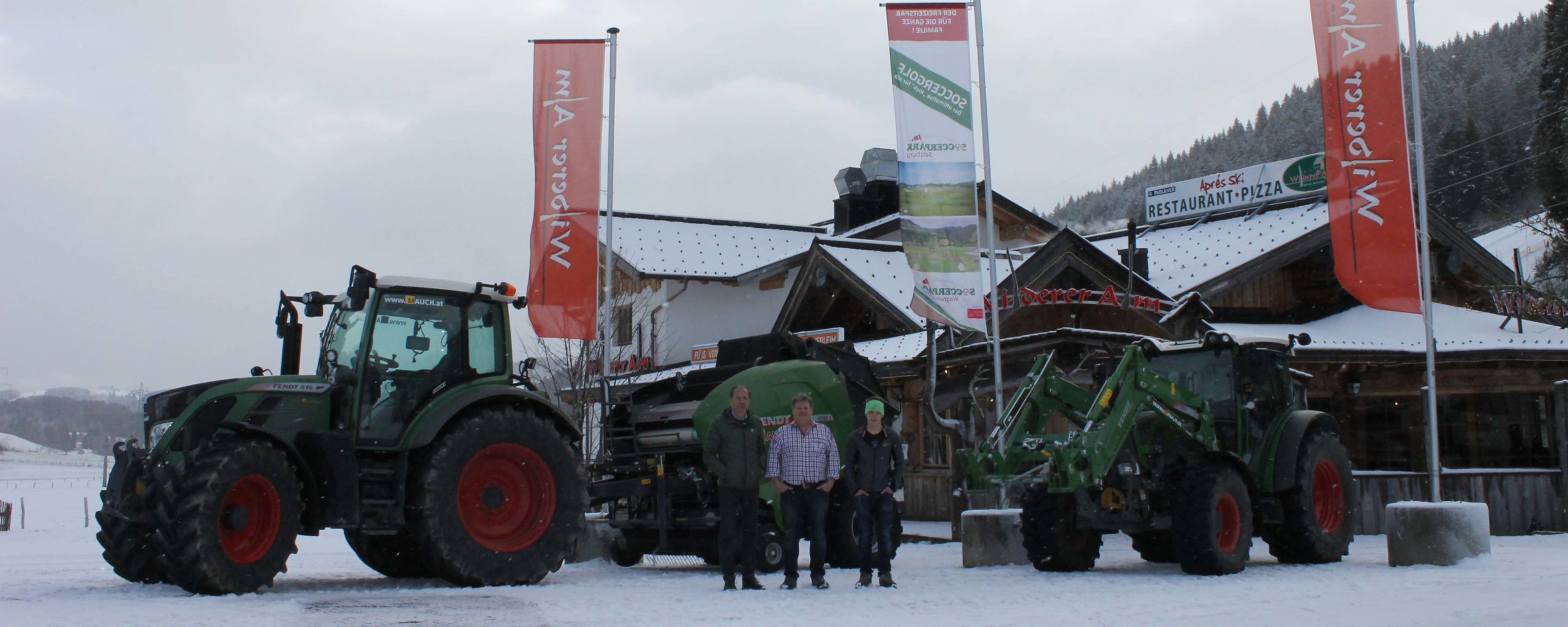 Drei Männer stehen zwischen zwei Fendt-Traktoren vor einem schneebedeckten Gebäude mit Fahnen von Mauch.
