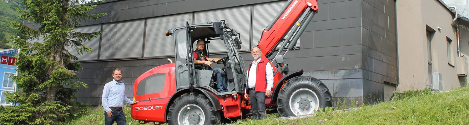 Drei Männer stehen um einen roten Weidemann-Lader neben einem Gebäude im Freien.