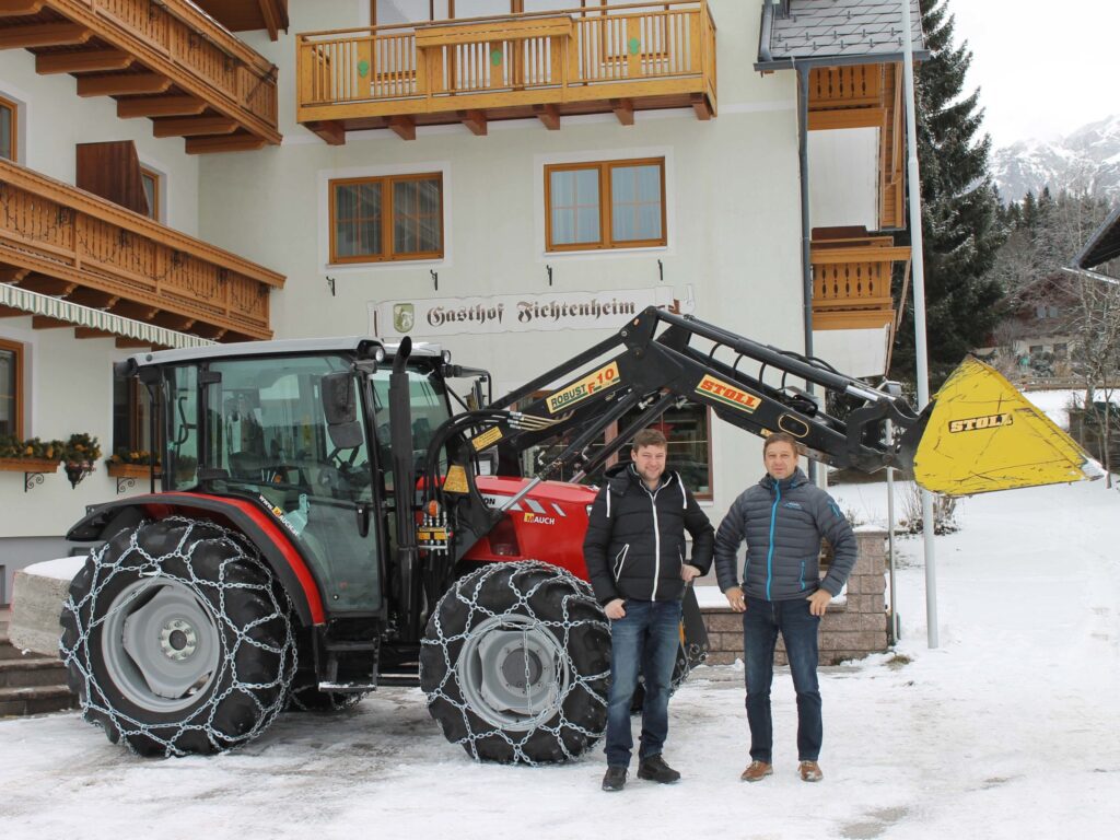 Zwei Männer stehen vor einem mit Schneeketten ausgerüsteten Traktor mit Stoll-Frontlader vor einem Gasthof.