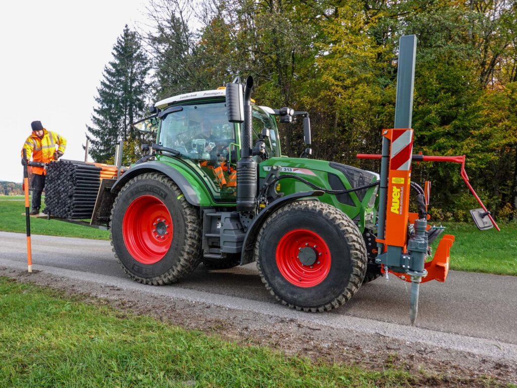 Un trattore Fendt 313 Vario con attrezzatura Auer e un operaio edile che erige un palo sul ciglio della strada.