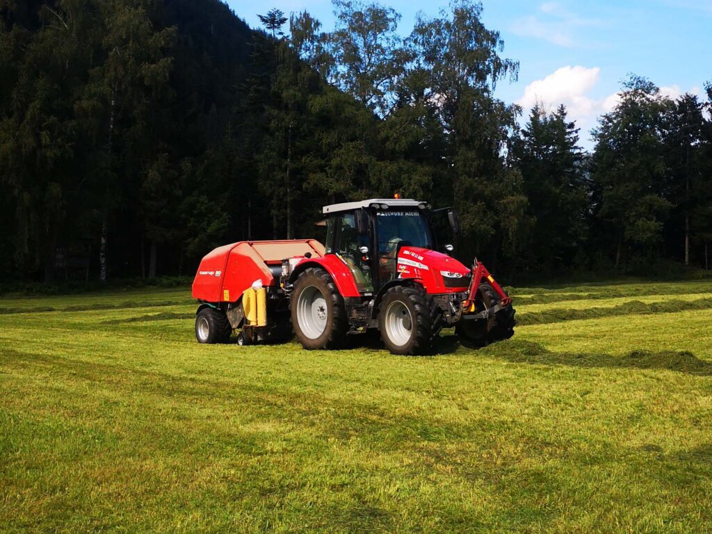 Traktor mit Ballenpresse auf einem Feld vor bewaldeter Kulisse.