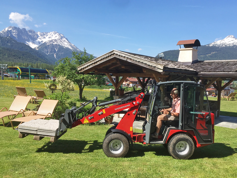 Ein Mann fährt einen Weidemann WM 1240LP Hoftrac mit einer geladenen Palette vor einer Berglandschaft.