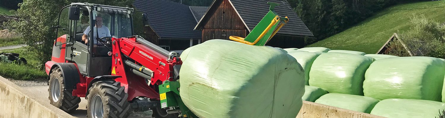 Ein Mann fährt einen roten Weidemann Radlader und transportiert eine große, grüne Heuballenrolle.