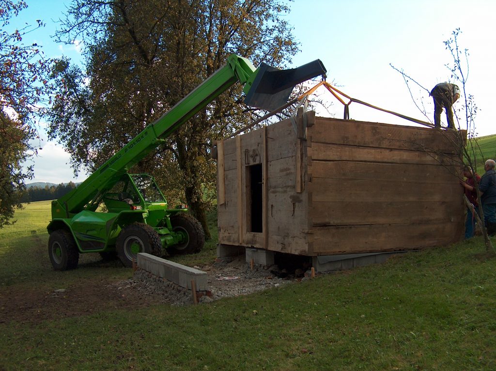 Teleskopski utovarivač podiže drvenu kolibu; dvije osobe stoje pokraj nje u prirodi.