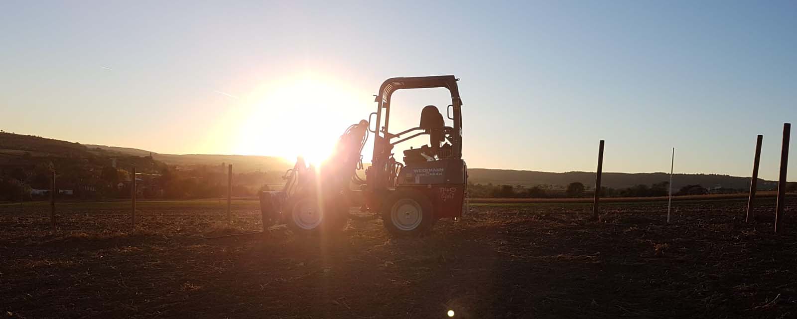 Ein Weidemann-Maschinenfahrzeug im Gegenlicht bei Sonnenuntergang auf einem Feld.