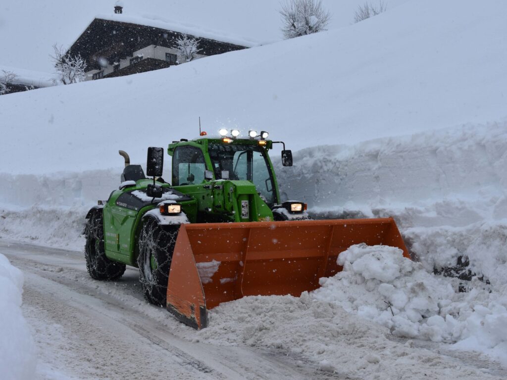 Зелени трактор са наранџастом лопатом за снег чисти снежну улицу испред куће.