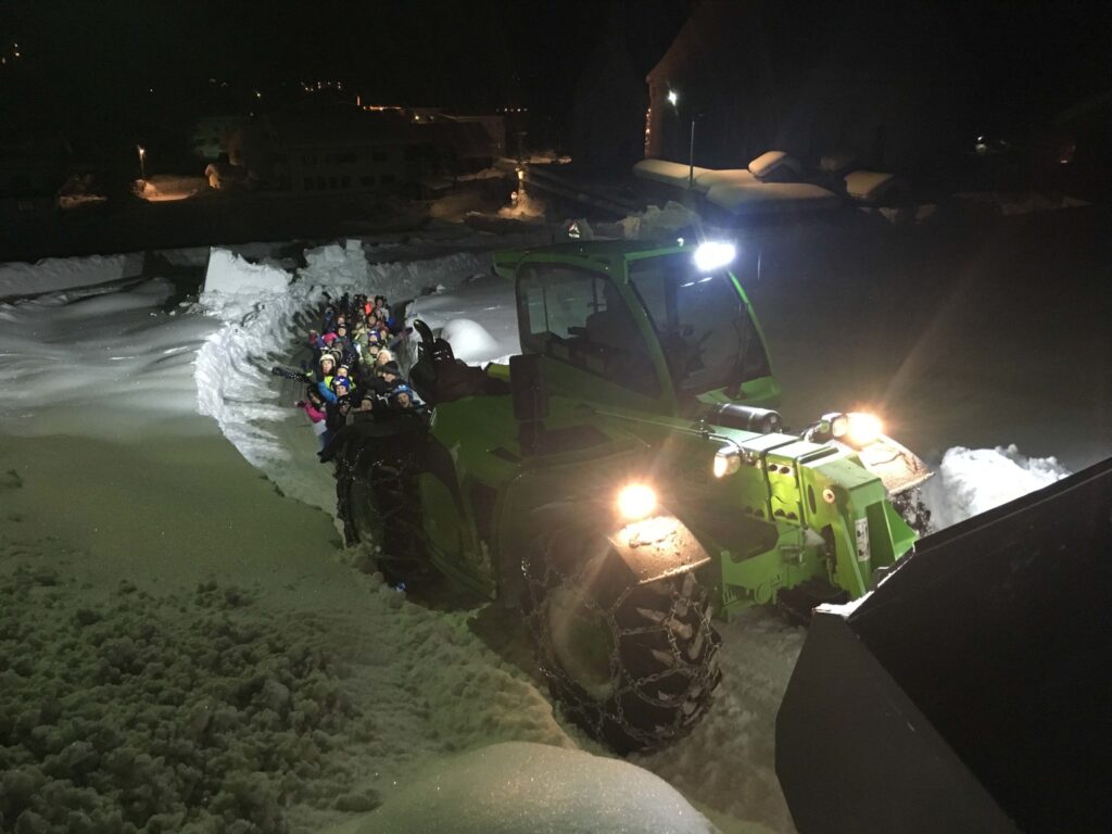 Zeleni bager utovarivač MAUCH noću gura skupinu nasmijane djece po snježnoj stazi.