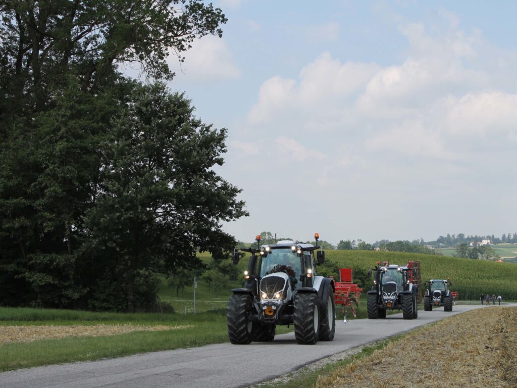 Tractoarele se deplasează unul în spatele celuilalt pe un drum printr-o zonă rurală.