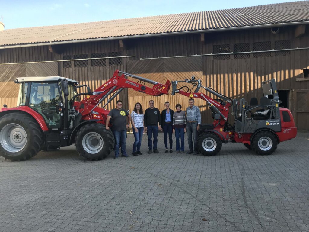 Eine Gruppe Menschen steht zwischen einem großen und einem kleinen Traktor vor einem Holzgebäude.