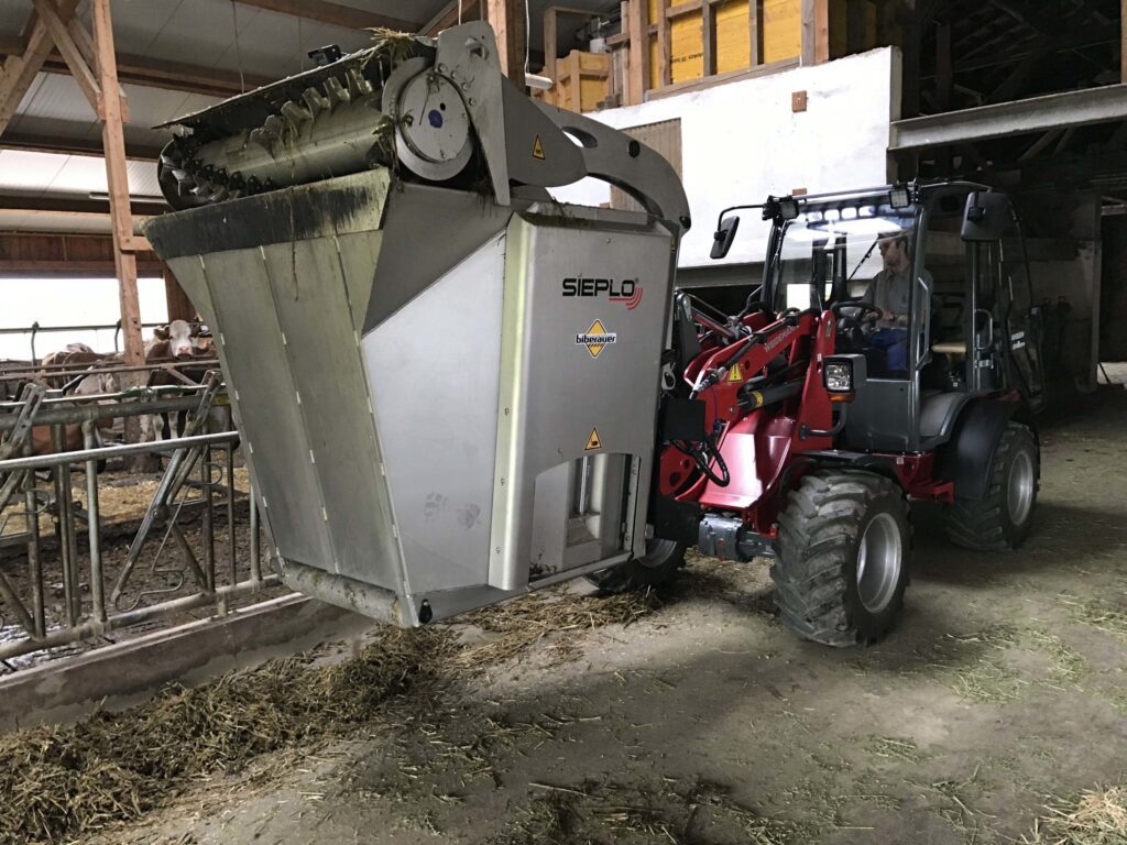 Ein roter Traktor befüllt mit einem Sieplo-MB1500-Anbaugerät ein Futtertrog in einem Stall.